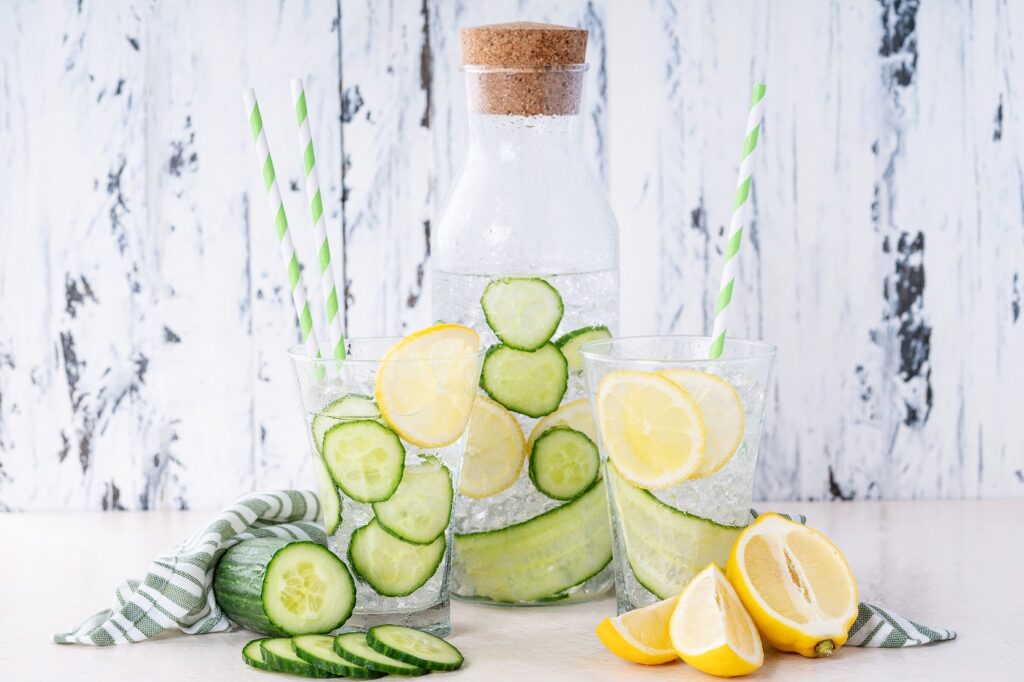 Cucumber and lemon infused water in glasses and bottle served with ice over bright white wooden background.